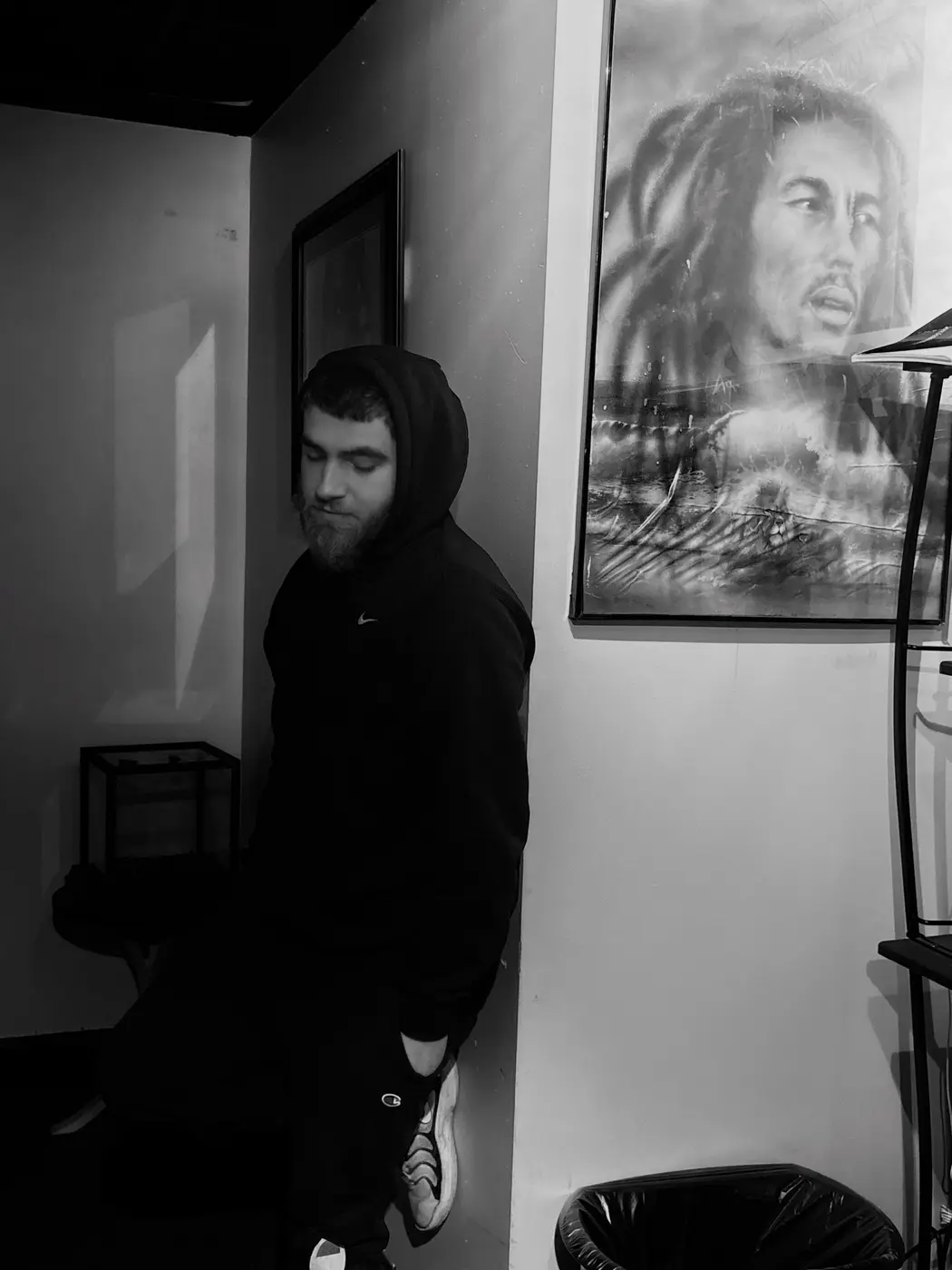 Michael Tennis Davis leaning on a wall beside a Bob Marley drawing, in black and white